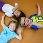 3 Kinder liegen mit dem Rücken auf dem Boden und winken mit ihren Armen