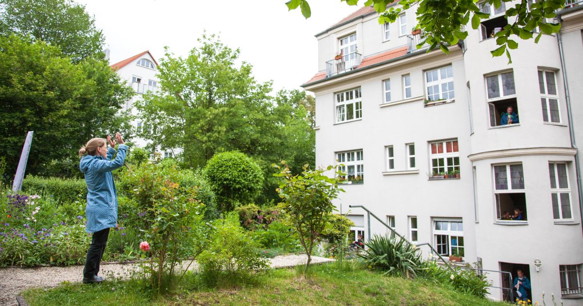 Eine Frau steht vor einem Wohnhaus in Jena und dirigiert die Bewohner, die aus dem Fenstern heraus ein Konzert geben