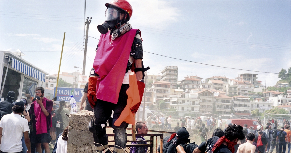 Fotografie von Julian Röder: Mit Gasmaske und Schutzbekleidung ausgestatteter Mann auf einer Barrikade mit Menschen und Häusern im Hintergrund