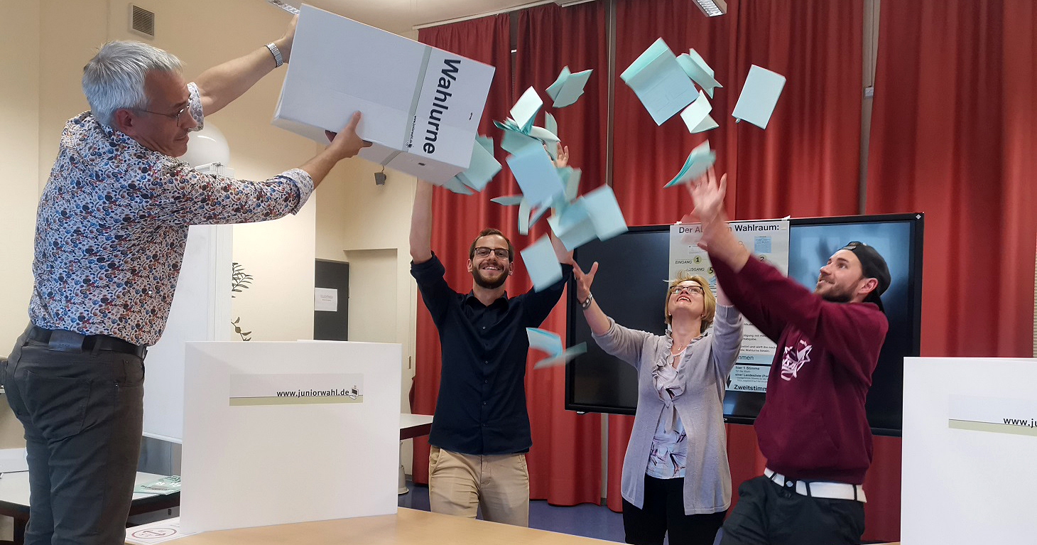 Das Team der vhs freut sich über die zahlreichen Stimmzettel bei der Juniorwahl 2021, vier Mitarbeiter:innen leeren die Wahlurne mit viel Vorfreude