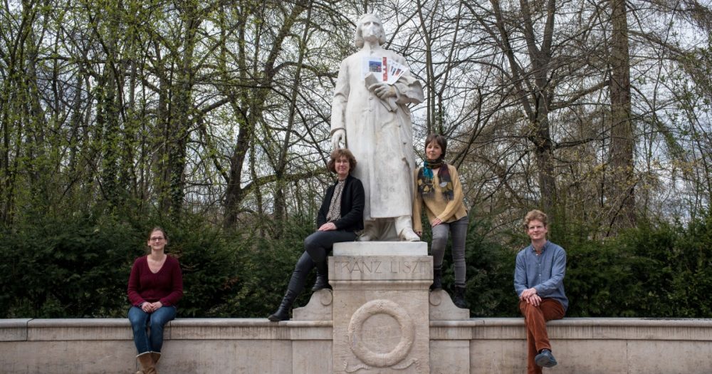 Das Redaktionsteam des SeitenKlangs an einer Franz Liszt-Statue, die eine Mund-Nase-Bedeckung trägt und Ausgaben des Magazins im Arm hält