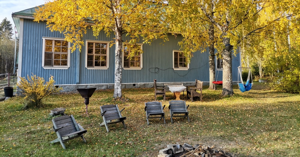 Feuerstelle mit Holzstühlen, im Hintergrund ein Holzhaus, weotere Gartenmöbel und herbstliche Birken