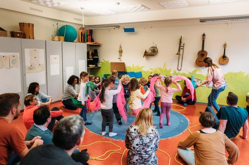viele kleine Kinder tanzen im Kreis mit bunten Tüchern zu Musik, drum herum sitzen die Eltern