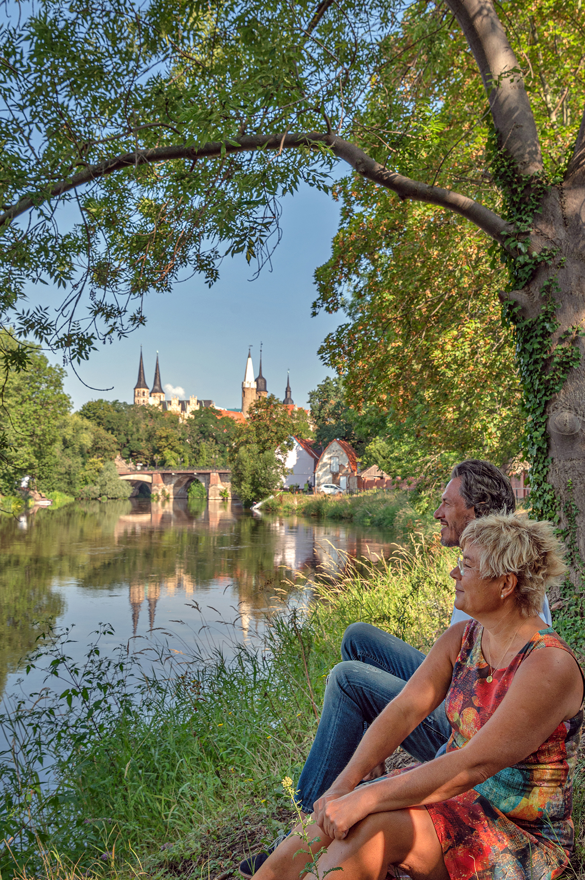 Ein Pärchen sitzt am Ufer der Saale und schaut auf Merseburg, im Hintergrund sieht am Dom und Schloss von Merseburg