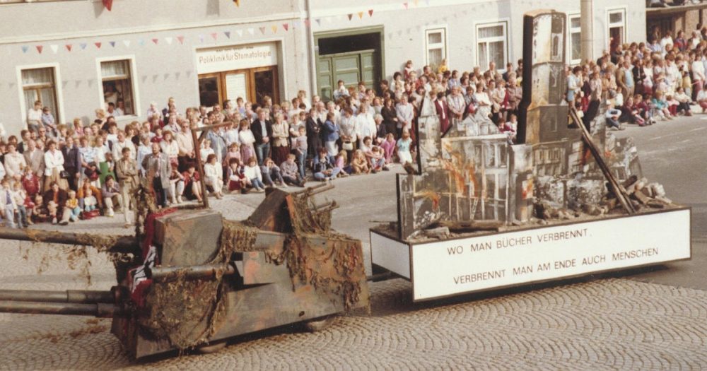 Fotografie eines Festumzugs in Jena von 1986, auf der ein als Panzer dekorierter Umzugswagen vor Publikum fährt, auf dessen Ladefläche eine brennende Stadt dargestellt wird mit der Aufschrift "Wo man Bücher verbrennt, verbrennt man am Ende auch Menschen"