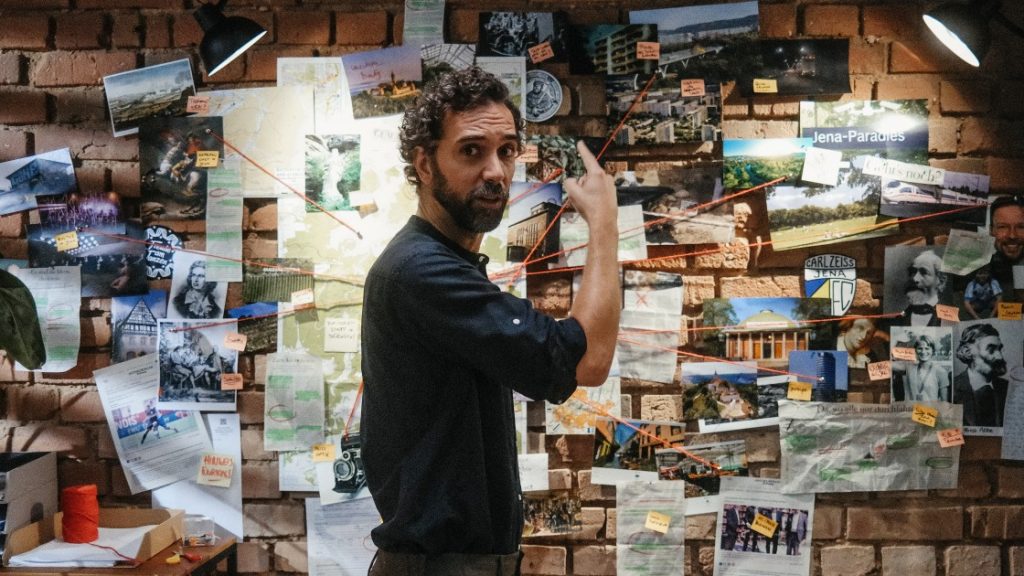 Ein Mann steht vor einer Pinnwand voller Bilder, Zeitungsartikel und Notizen, die mit einem roten Faden verbunden sind