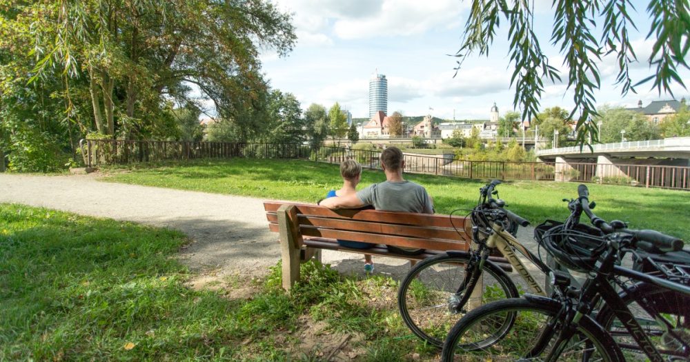 Paar sitzt mit Fahrrädern auf einer Parkbank an der Saale und blickt auf die Jenaer Innenstadt