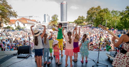 Kulturarena Jena, Kinder stehen auf der Bühne, davor ein voller Zuschauerraum Open Air und ein Blick auf den Jentower