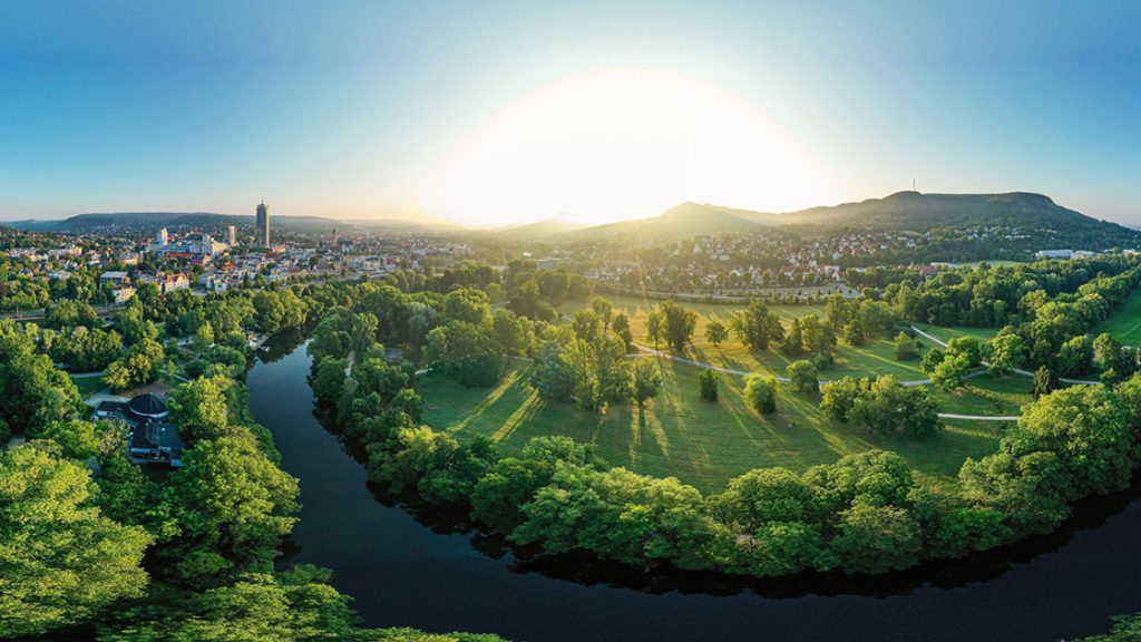 Blick auf Jena, von oben, ins grüne, auf die Saale, im Hintergrund die Berge und links die Lichtstadt Jena, die Sonne geht gerade auf