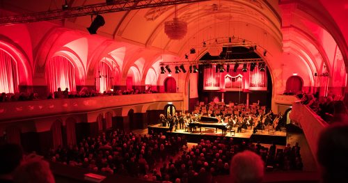 Jenaer Philharmonie im Volkshaus Jena, der Saal ist rot beleuchtet und voller Menschen, auf der Bühne spielen die Musiker:innen