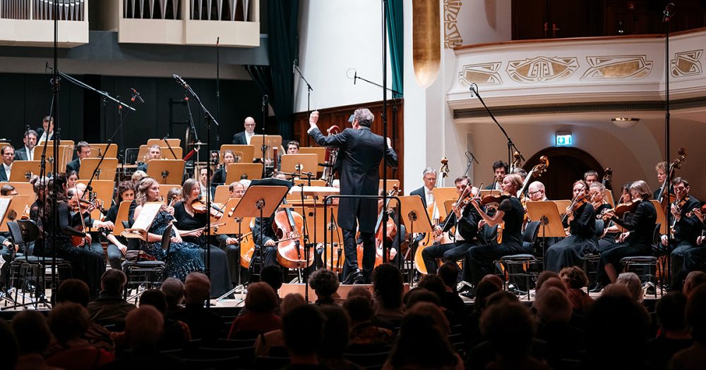 Das Volkshaus Jena mit vollem Publikum, Blick auf die Bühne, auf der die Jenaer Philharmonie spielt und Simon Gaudenz dirigiert