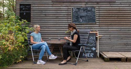 Das Salü in Jena, von außen, 2 Frauen sitzen vor einer Hiolzwand und trinken Wasser im Außenbereich
