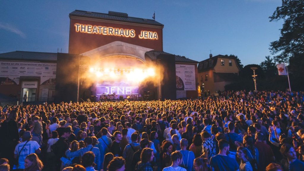 Publikum bei der Kulturarena vor der hell erleuchteten Bühne des Theaterhauses Jena, auf der in Lichtschrift "Jena" steht