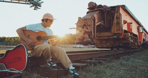 Der Musiker Bosse mit einer Akustikgitarre auf einer Bahnschiene sitzend, neben ihm steht ein Bauzug, über seinem Kopf ist ein Kran zu sehen