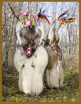 Fotografie von Charles Fréger aus der Serie "Wilder Mann": Zwei Menschen stehen zwischen Laubbäumen, sie tragen dricke, langhaarige Pelzkostüme und auch auf dem Kopf riesige Pelze, die ihre Gesichter verdecken. Darauf sind Geweihe und bunte Stoffbänder befestigt.