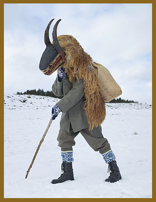 Fotografie von Charles Fréger aus der Serie "Wilder Mann": Mensch geht an einem Spazierstock durch eine Schneelandschaft, er trägt einen braunen Pelz auf dem Rücken, darauf einen Jutesack, und eine Maske, die sich zu einer Schnauze mit spitzen Zähnen wölbt und Hörner auf dem Kopf.