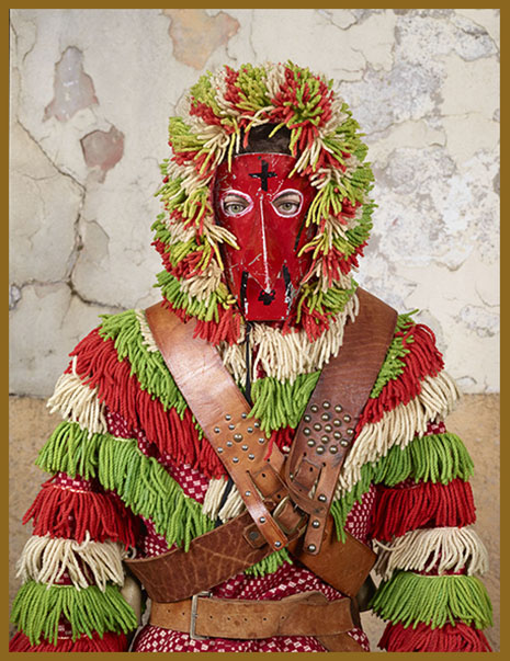 Fotografie von Charles Fréger aus der Serie "Wilder Mann": Brustportrait eines Menschen in einem Kostüm aus grünen, beigen und roten Wollfäden, mit einer roten Maske, die eine lange, spitze Nase formt und braunen Gürteln, die über Brust und Taille geschnürt sind.