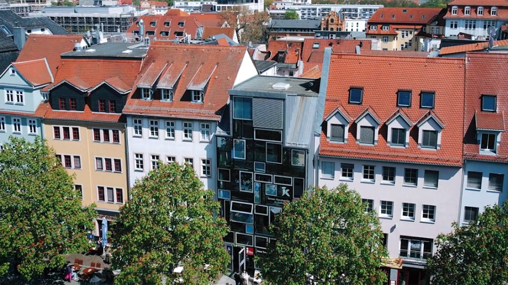 Blick von oben auf die Jena Tourist-Information am Markt 16 im Herzen Jenas, ringsrum Häuser, die moderne Glasfassade, davor Kastanienbäume