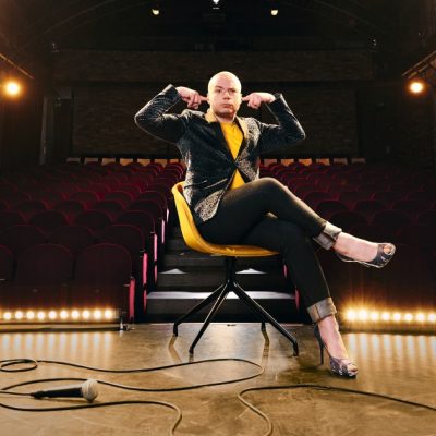 Comedian Okan Seese in Highheels auf einem Stuhl in einem Theatersaal hält sich die Finger in die Ohren
