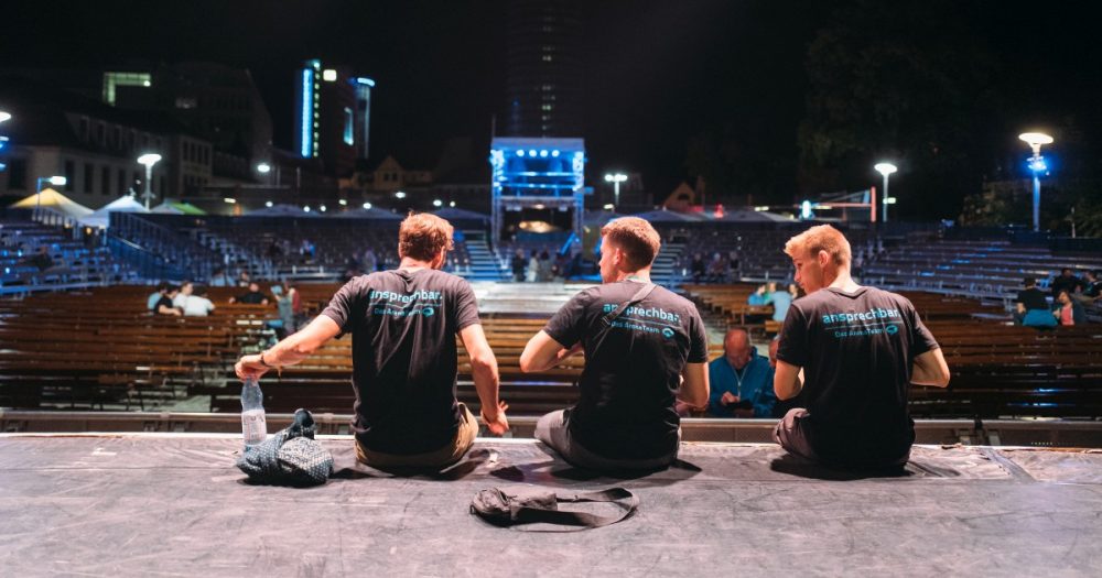 Drei Mitglieder des Kulturarena-Teams mit T-Shirts, auf denen "ansprechbar" steht, sitzen nebeneinander auf der Bühne der Kulturarena nach einem Konzert