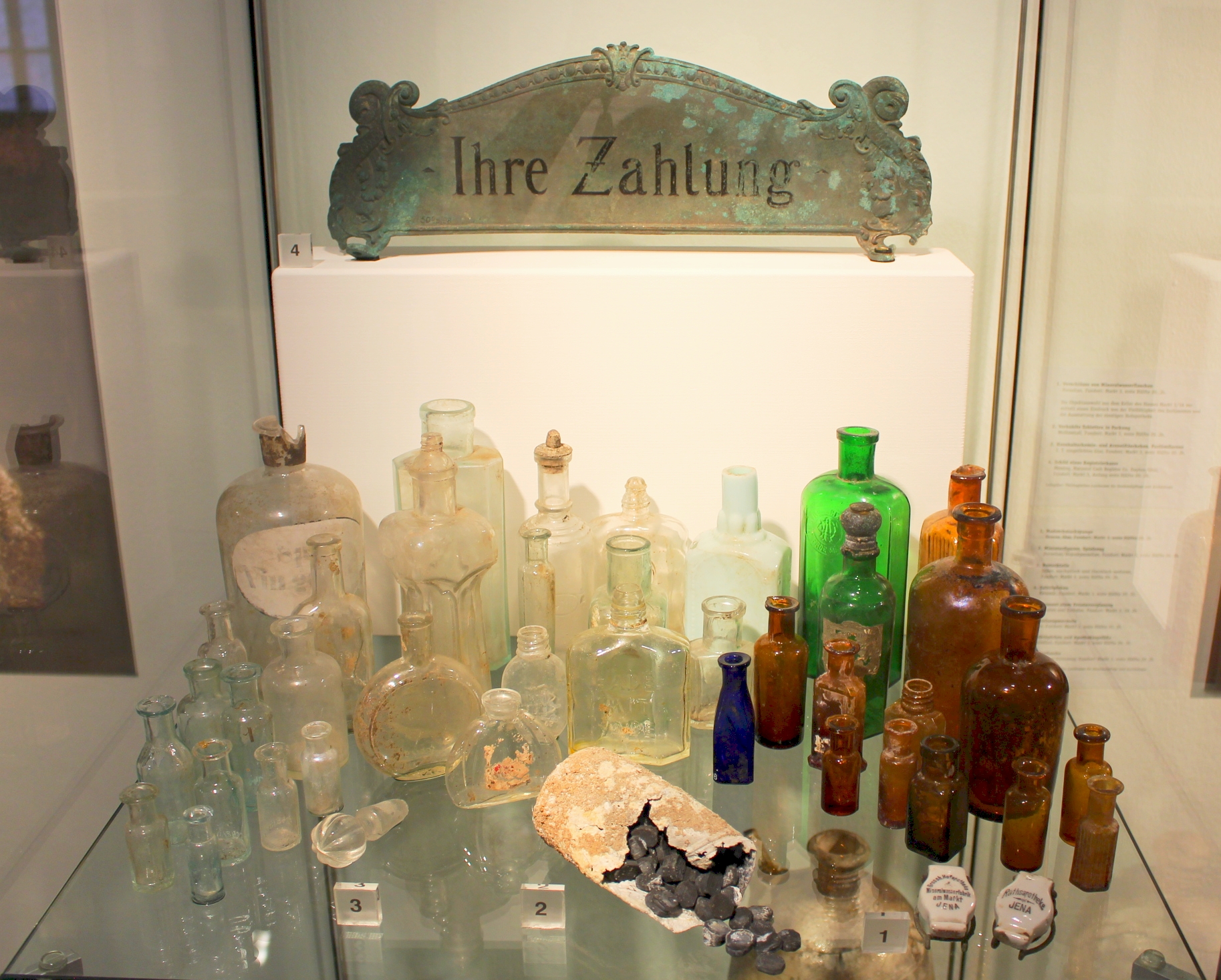 Blick in die Ausstellung: Glasgefäße, Keramik, Schild einer Registrierkasse und Pastillen aus der ehemaligen Hofapotheke, Mitte 20. Jahrhundert