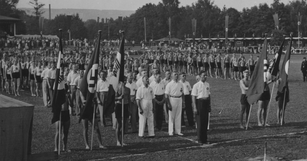 Fahnenappell des Nationalsozialistischen Reichsbundes für Leibesübungen, im Hintergrund Sportler:innen der Sprung- und Laufwettbewerbe
