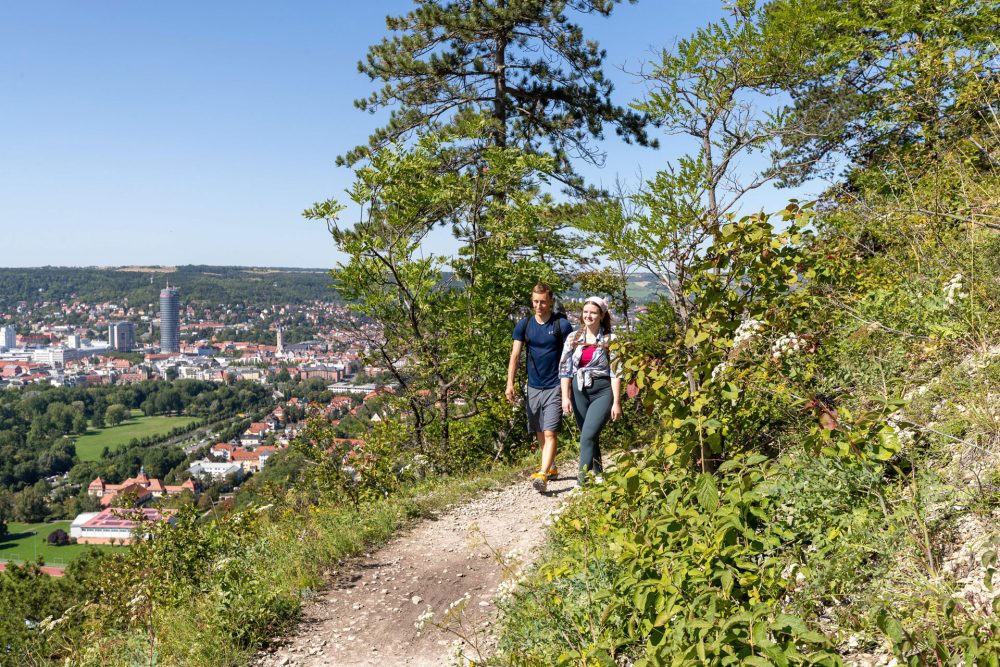 Ein Pärchen wandert mit Rucksäcken die SaaleHorizontale an einem Hang entlang, der Weg bietet eine tolle Sicht auf die Stadt Jena