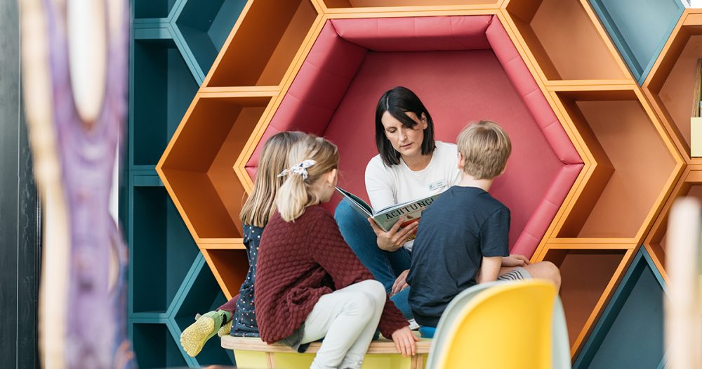 Bibliothekarin hockt in einer gepolsterten, bunten, wabenförmigen Wand in der neuen Kinderabteilung der Ernst-Abbe-Bücherei Jena und liest drei Kindern ein Buch vor