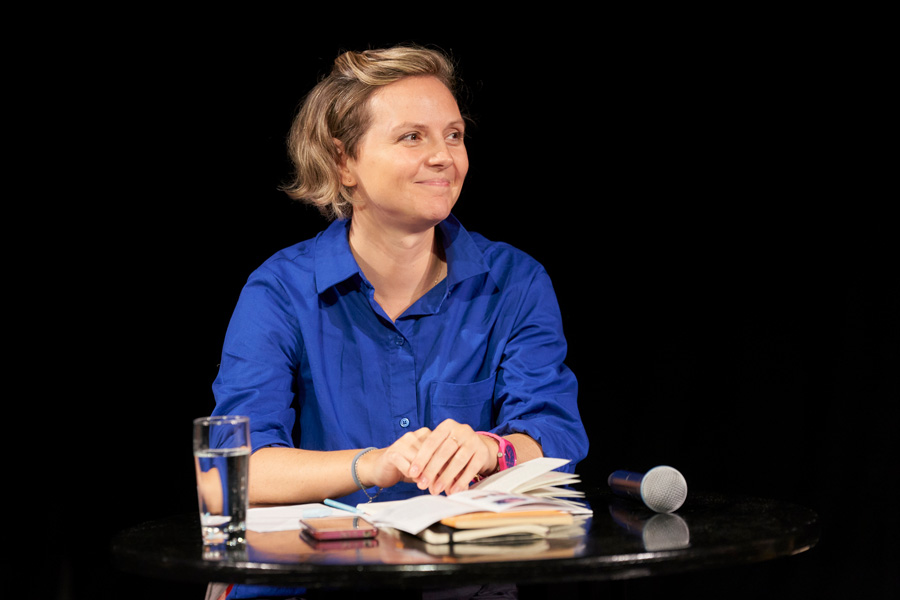 Eine Frau mit kürzerem blonden Haar und einer royalblauen Blus sitzt an einem Tisch, auf dem ein Wasserglas, Notizen, ein Buch und ein Mikrofon liegen