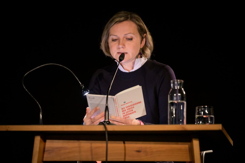 Eine Frau mit kürzerem blonden Haar, die vor einem schwarzen Hintergrund an einem Mikrofon sitzt, liest aus dem Buch „Die Verteidigung der Poesie in Zeiten dauernden Exils”