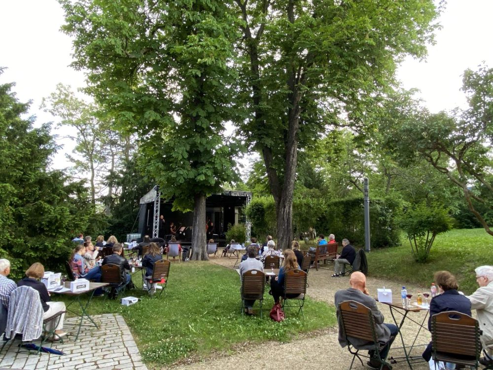 Im Garten der Villa Rosenthal stehe eine Bühne, Tisch und Stühle, an denen Gäste einem Konzert lauschen.