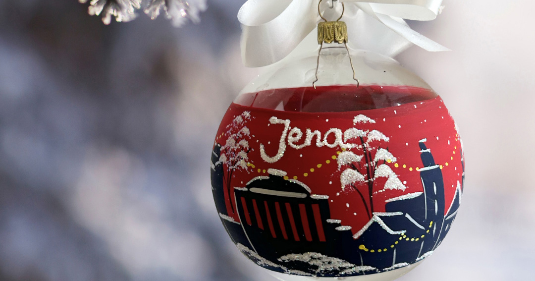 Mit der Skyline der Stadt Jena bemalte rote Weihnachtskugel, welche an einem frostigen Tannenzweig hängt.