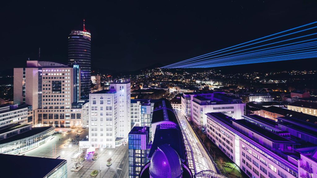 Vogelperspektive auf das Jenaer Stadtzentrum mit Unicampus, Jentower, Zeiss-Hochhaus und am Horizont Laserstrahlen, die in den Himmel ragen