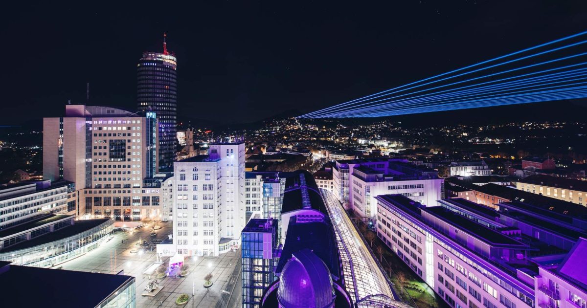 Vogelperspektive auf das Jenaer Stadtzentrum mit Unicampus, Jentower, Zeiss-Hochhaus und am Horizont Laserstrahlen, die in den Himmel ragen