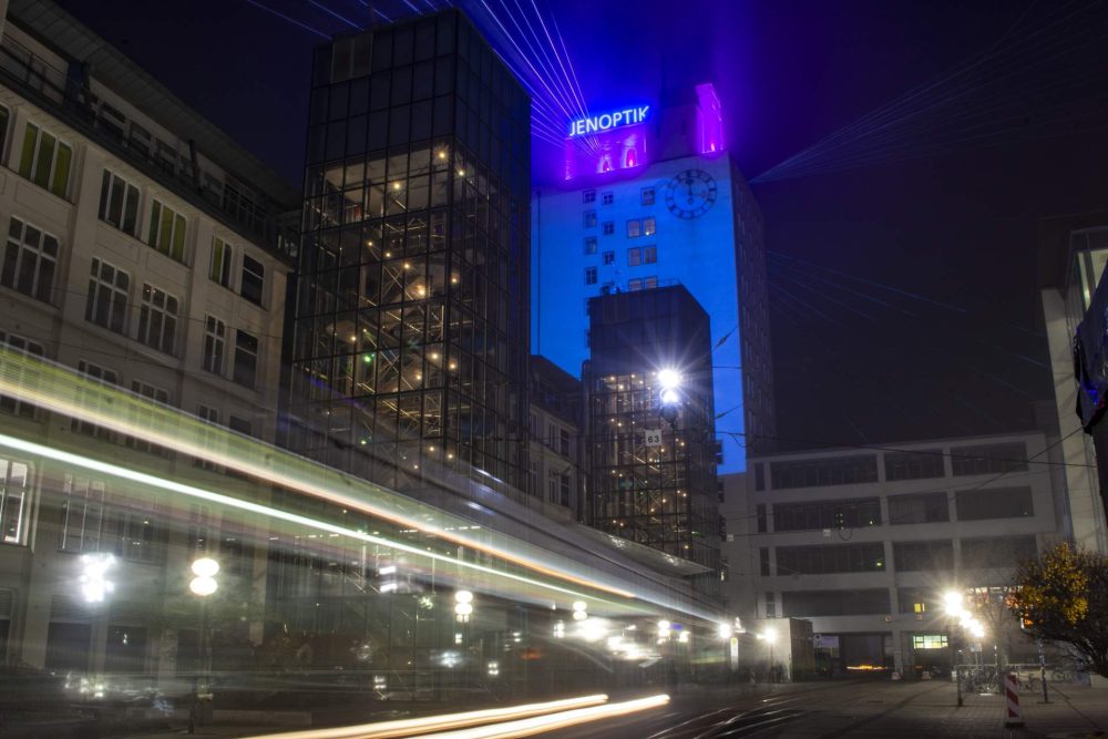 blau-beleuchteter Jenoptik-Tower mit Schriftzug des Unternehmens mit Blick auf den Unicampus und Lichtern einer vorbeifahrenden Straßenbahn