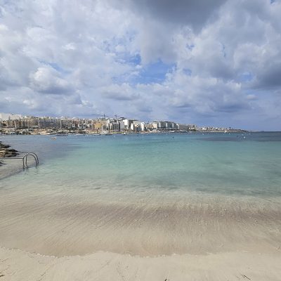 Ein Strand in Malta, das Meer ist grün-blau, dahinter türmt sich eine Stadt auf