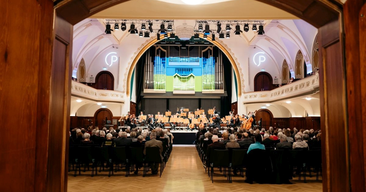 Blick durch die Flügeltüren in den großes Saal des Volkshauses Jena: Publikum, Jenaer Philharmonie auf der Bühne, die Orgel darüber wird in den Ukrainischen Nationalfarben angestrahlt