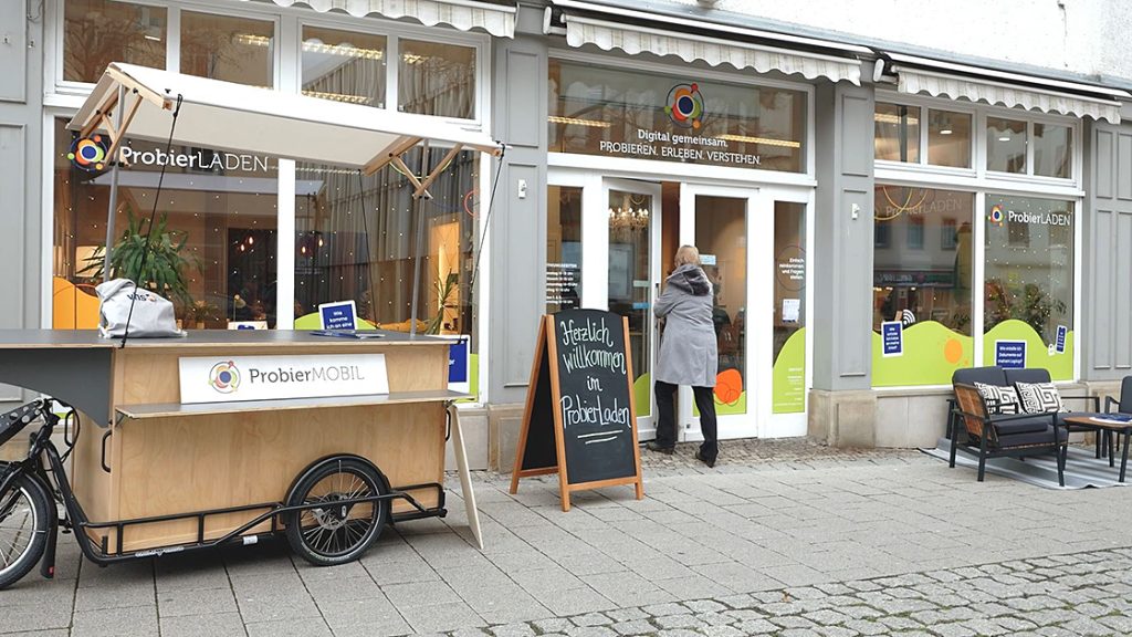 Frau betritt den ProbierLaden Jena, davor steht das ProbierMobil und ein Willkommen-Aufsteller