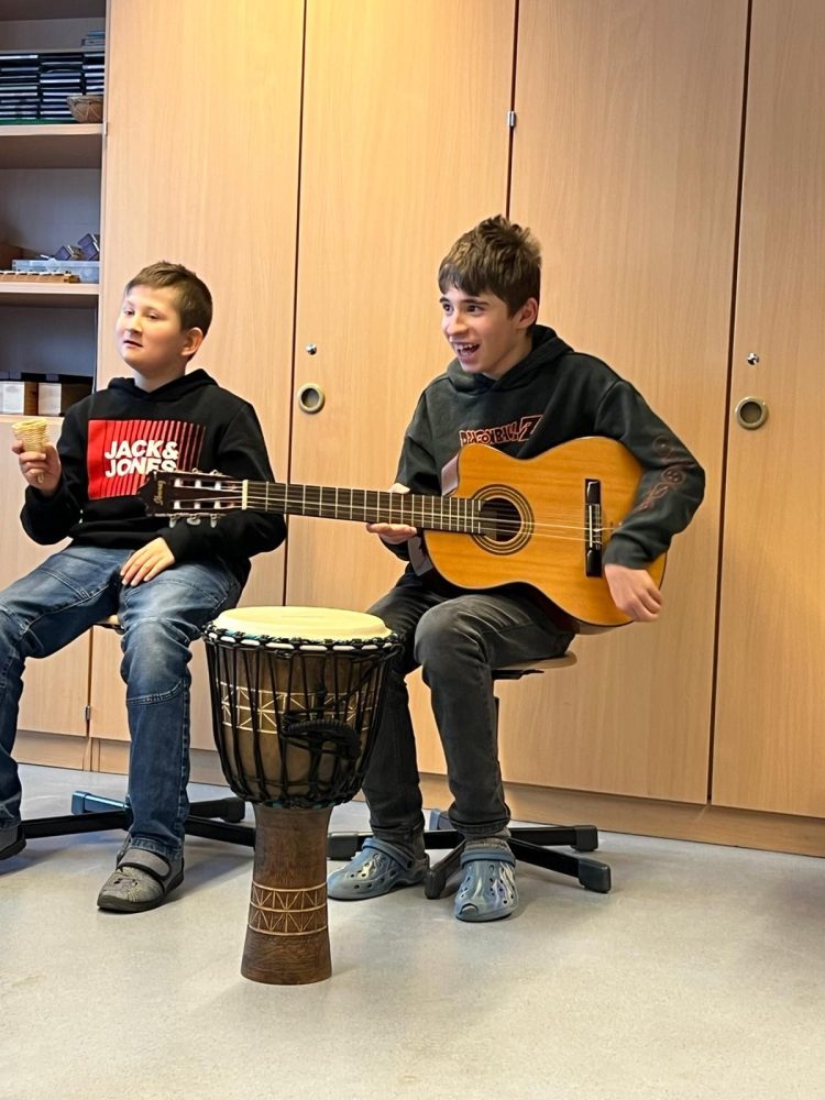 Zwei Kinder sitzen nebeneinander und musizieren gemeinsam. Ein Kind hat eine Gitarre auf dem Schoß liegen. Das andere Kind hält ein kleines Instrument in der Hand. Beide haben einen fröhlichen Gesichtsausdruck.