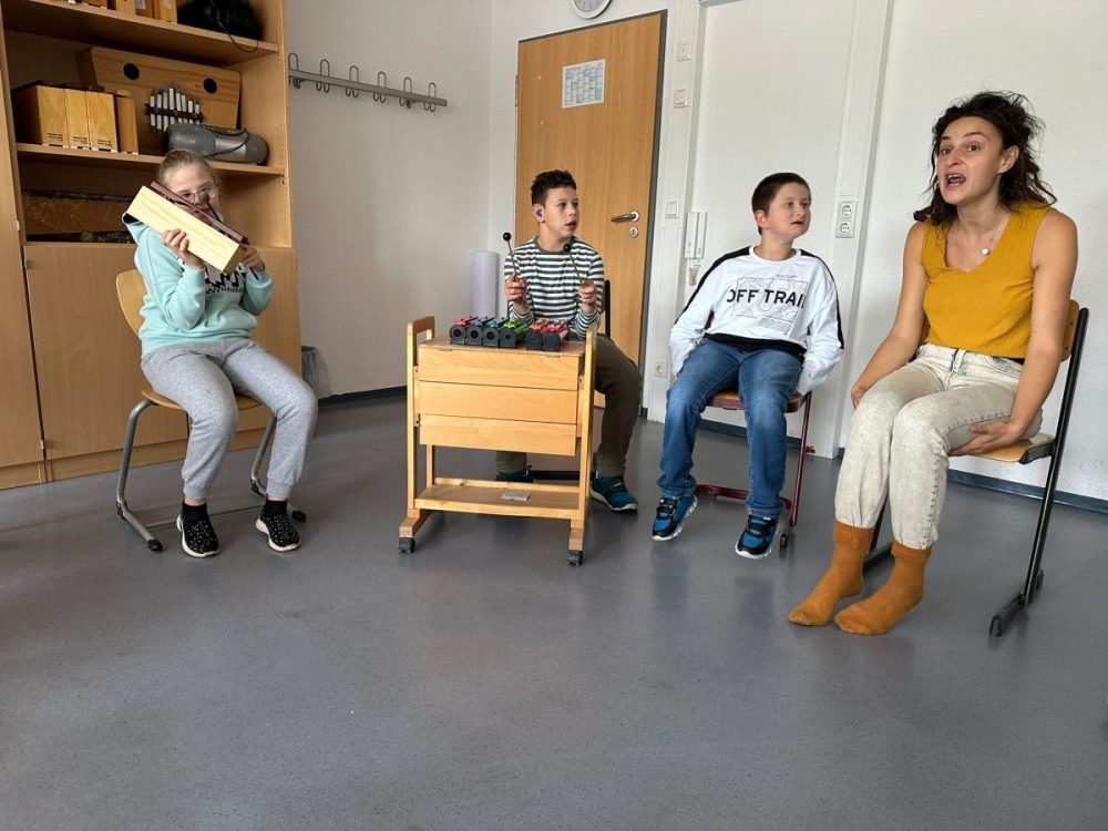 3 Kinder musizieren gemeinsam mit der Musischullehrerin. Die Lehrerin sitzt ganz rechts in der Reihe und hat einen offenen Mund als ob sie gerade singt. Das Kind neben ihr schaut sie an und hat ebenfalls den Mund geöffnet. Das dritte Kind sitzt vor eiem bunten Xylophon und hat 2 Schlägel in der Hand. Das dritte Kind in der Reihe hat ebenfalls ein Schlaginstrument und Schlägel in der Hand.