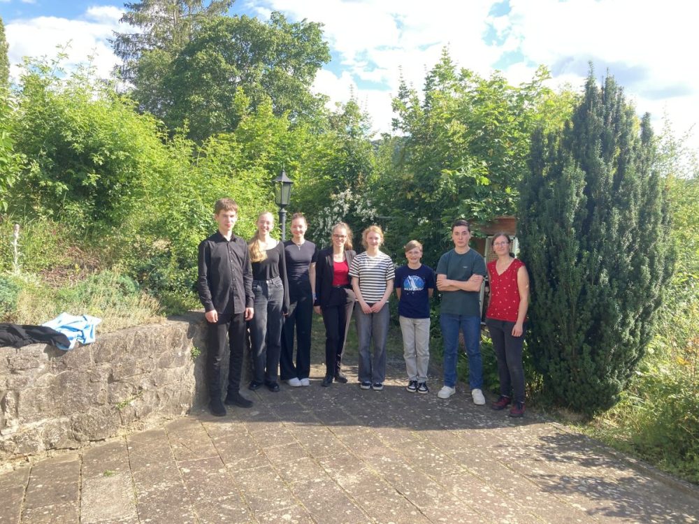 Das Foto zeigt eine Gruppenaufnahme aller Mitwirkenden des 1. Kurzkonzertes – diesmal ohne Instrumente. Die Gruppe steht am Rand der Terrasse. Im Hintergrund sind Bäume und Büsche zu sehen.