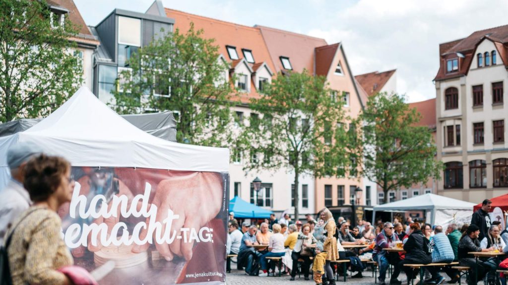 Zwei Gäste der handgemacht-Tage laufen über den Jenaer Marktplatz und kommen dabei an einem Pavillon vorbei, der den Namen der Veranstaltung trägt, im Hintergrund befinden sich weitere auf Bierbänken sitzende Gäste.