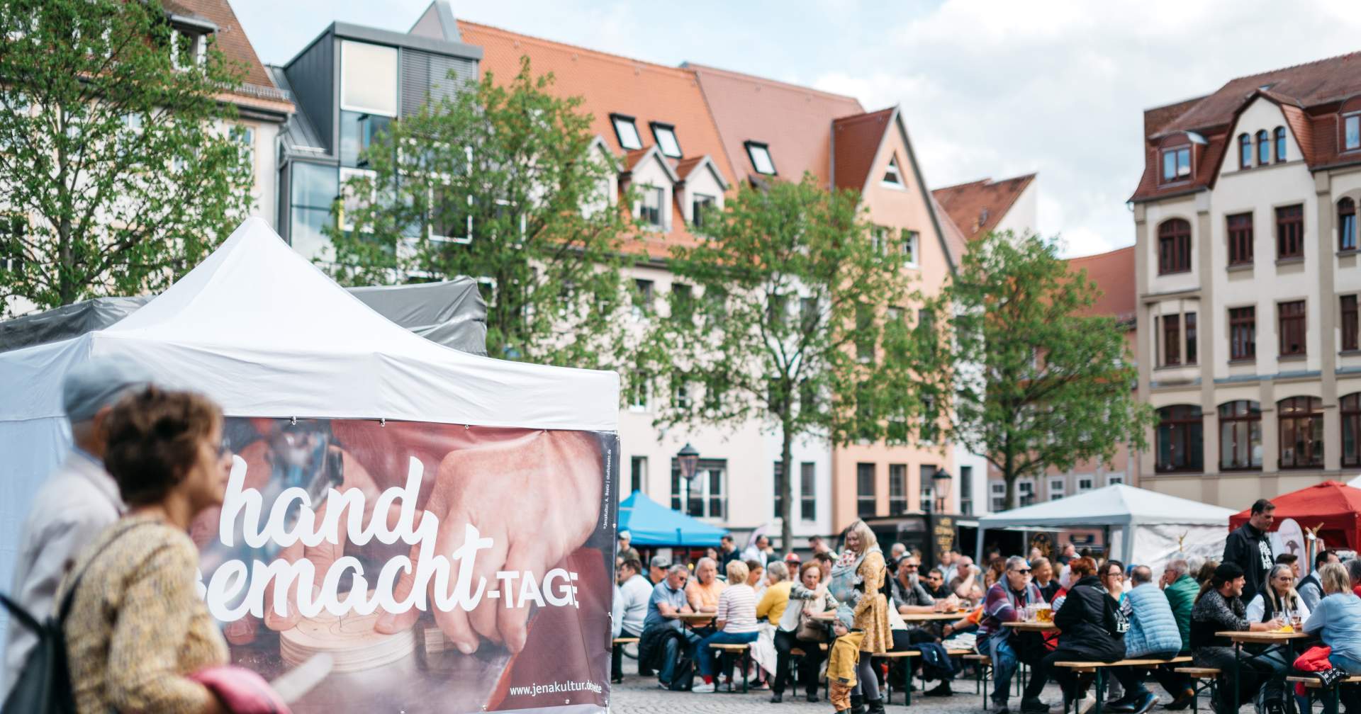 Zwei Gäste der handgemacht-Tage laufen über den Jenaer Marktplatz und kommen dabei an einem Pavillon vorbei, der den Namen der Veranstaltung trägt, im Hintergrund befinden sich weitere auf Bierbänken sitzende Gäste.