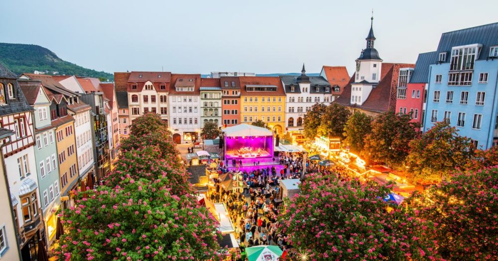 Der Frühlingsmarkt von oben in Blickrichtung Bühne. Personen stehen auf dem Marktlatz verteilt übrall stehen Essens und Getränkebuden. Die Bäume auf dem Frühlingsmarkt blühen. Auf der Bühne spielt gerade eine Band.