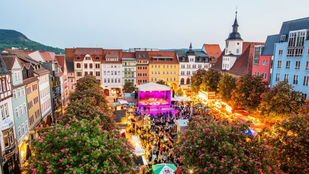 Der Frühlingsmarkt von oben in Blickrichtung Bühne. Personen stehen auf dem Marktlatz verteilt übrall stehen Essens und Getränkebuden. Die Bäume auf dem Frühlingsmarkt blühen. Auf der Bühne spielt gerade eine Band.