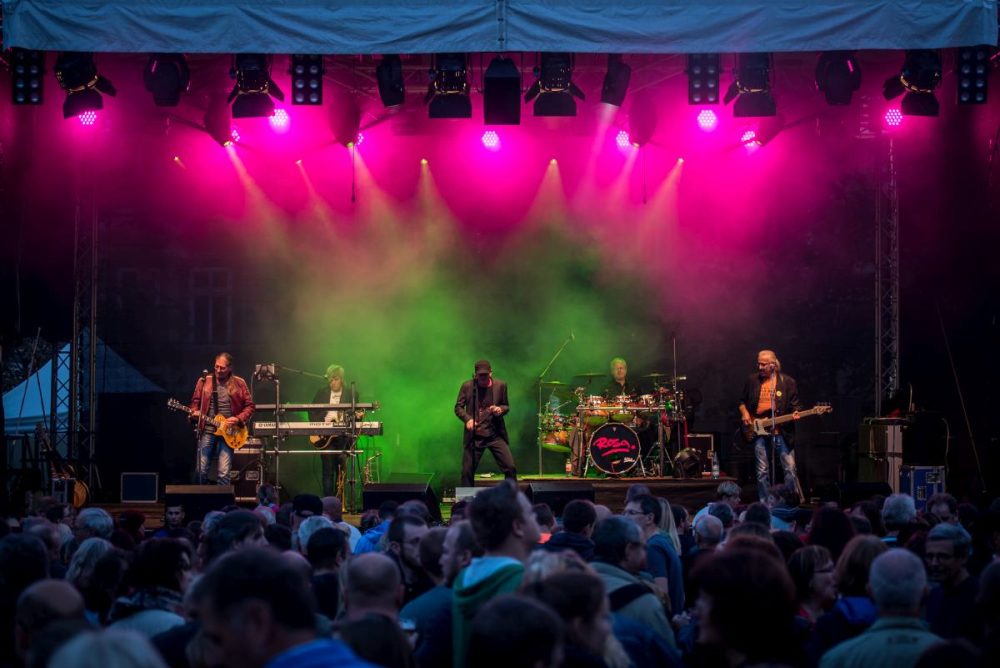 Die Bühne auf dem Jenaer Altstadtfeste. Auf der Bühne stehen 5 Personen, die zu einer Band gehören. Eine Person steht links auf der Bühne und hat eine Gitarre in der Hand. Eine Person steht rechts auf der Bühne und hat eine Bassgitarre in der Hand. In der Mitte der Bühne steht eine Person mit Mikro in der Hand. Hinten links steht eine Person am Keyboard und hat eine Gitrarre um. Hinten rechts sitzt eine Person am Schlagzeug. Vor der Bühne stehen Personen, die zu schauen.