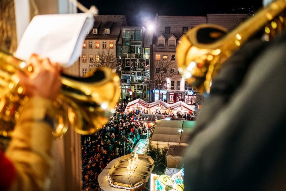 zwei Posaunen auf dem Jenaer Weihnachtsmarkt beim Turmblasen mit Blick Richtung Jena Tourist Information, davor stehen Buden und eine Bühne und Personen, die den Weihnachtsmarkt besuchen.