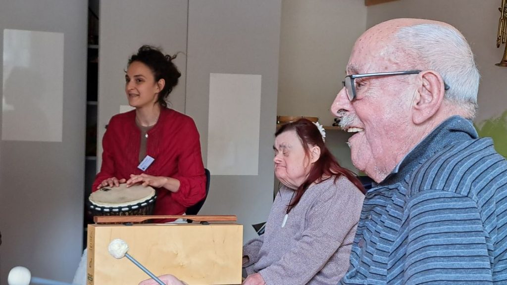 Rechts im Vordergrund des Bildes musiziert ein älterer Herr auf dem Metallophon und lächelt dabei. Im Hintergrund sieht man die Musikpädagogin Besjana Cegrani (links) an der Djembe und eine Teilnehmerin des Projekts am Klangbaustein.