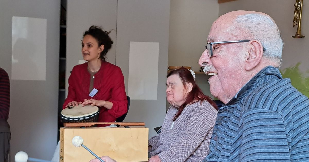 Rechts im Vordergrund des Bildes musiziert ein älterer Herr auf dem Metallophon und lächelt dabei. Im Hintergrund sieht man die Musikpädagogin Besjana Cegrani (links) an der Djembe und eine Teilnehmerin des Projekts am Klangbaustein.