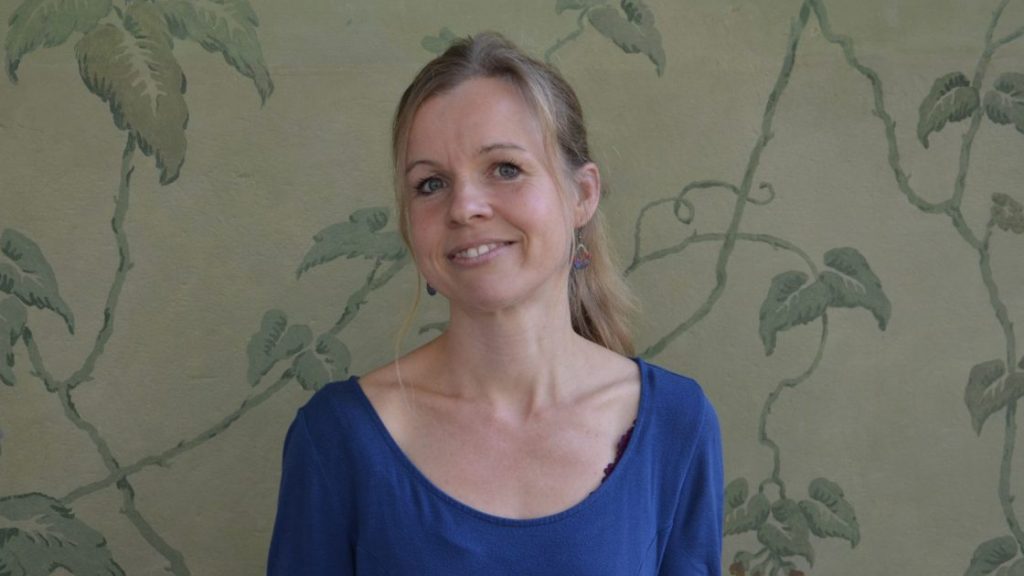 Eine blonde Frau mit zurückgebundenen Haar und blauem Shirt steht vor einer grünlichen Mustertapete.